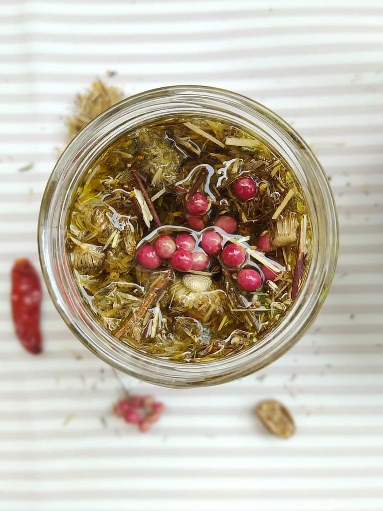 Crear extractos vegetales efectivos para tus fórmulas cosméticas 1 Oleato casero en tarro de vidrio visto desde arriba, con flores secas y pimienta rosa macerando en aceite